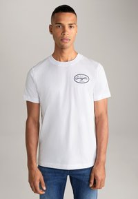 Weißes Baumwoll-T-Shirt mit rundem Halsausschnitt, kurzen Ärmeln und einem navyblauen Logo-Patch auf der linken Brustseite. Schlichtes Design, lässige Passform.