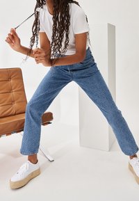 Blå mönstrade jeans med rak passform, bärs med en vit t-shirt och vita platåskor, uppställda bredvid en brun stol.