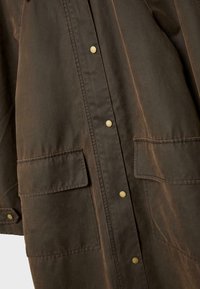 Giacca marrone scuro con due grandi tasche anteriori a pattina e bottoni a pressione in ottone lungo la chiusura centrale.