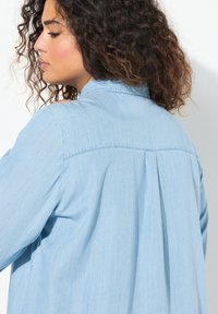 Camicia azzurra chiara, simile al denim, con una vestibilità comoda, maniche lunghe, colletto curvo e una piega sul retro per ulteriore texture.