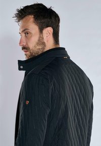 Black quilted jacket with a high collar, featuring a zip closure, diagonal stitching, and an orange accent near the collar.