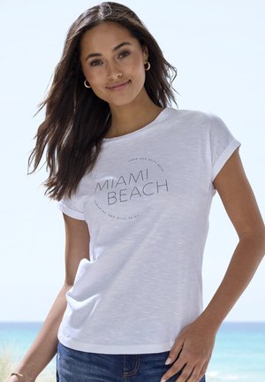 White cotton t-shirt with rolled sleeves featuring "MIAMI BEACH" text design and circular accents, paired with blue denim jeans.