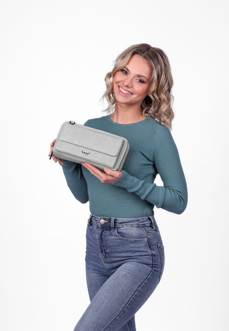 Usměvavá žena s vlnitými blond vlasy, která má na sobě tyrkysový svetr a džíny, drží světle šedou obdélníkovou kabelku na zip s klopou.