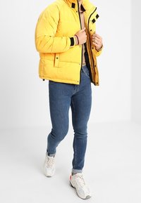 Žlutá péřová bunda s černými detaily, zapínání na zip a vycpaný design. Nositá přes šedé tričko, v kombinaci s modrými skinny džíny a teniskami.