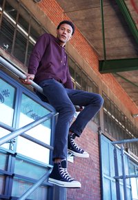 Bordeauxrote Kapuzenjacke, dunkle Jeans und schwarze High-Top-Sneaker mit weißen Sohlen. Model sitzt auf Gerüst, im Hintergrund eine Ziegelmauer und Fenster.