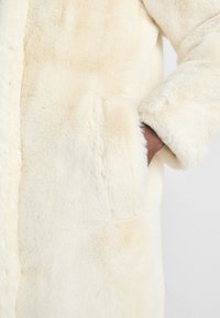 Kræmfarvet faux fur-frakke med en blød tekstur, der har sidelommer og en lige silhuet. Stoffet har en glat, blød overflade.