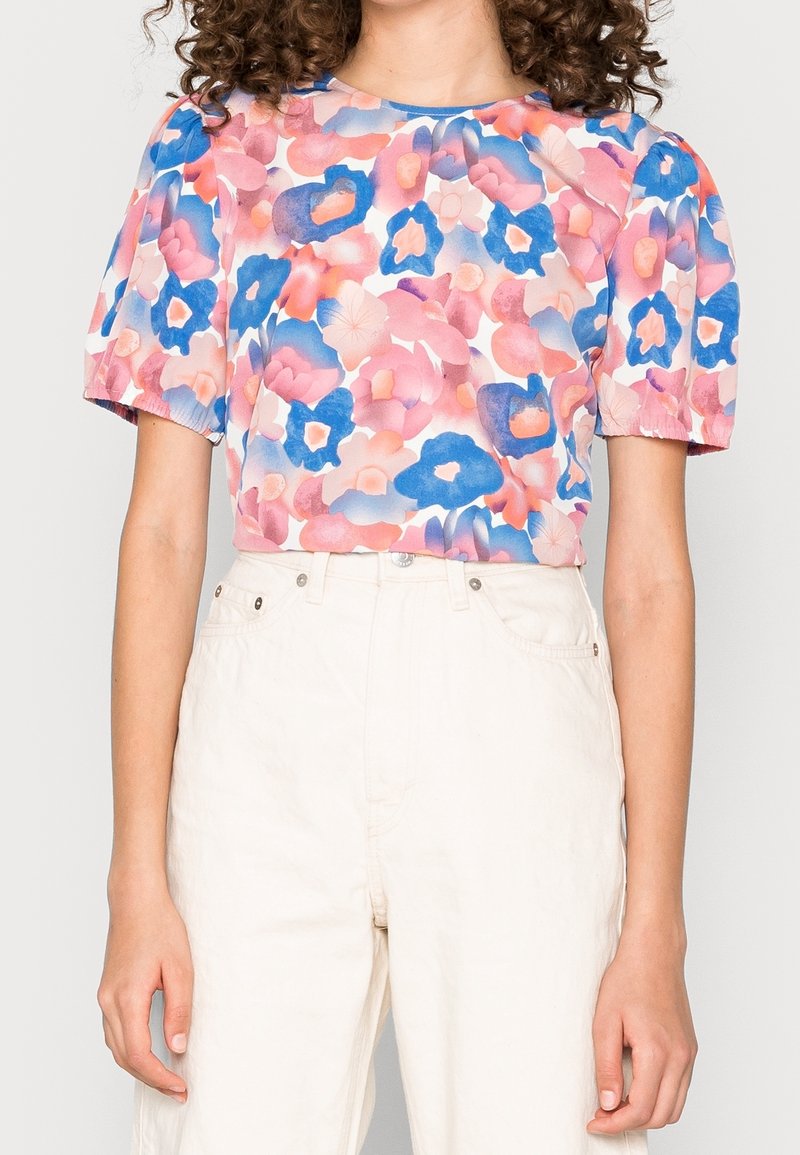 Femme portant un chemisier à motif floral rose, bleu et pêche avec de courtes manches bouffantes, associé à un pantalon taille haute blanc cassé.