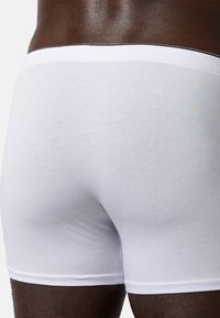 Pantaloni boxer bianchi in cotone con una fascia in vita nera, design aderente e texture liscia, mostrati da un'angolazione posteriore su pelle scura.