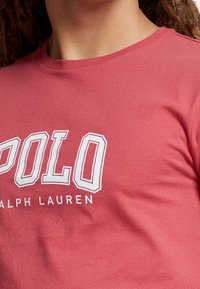 Röd bomulls t-shirt med rund halsringning som har stor vit text "POLO" och "RALPH LAUREN" i mindre teckenstorlek under. Slät textur, avslappnad design.