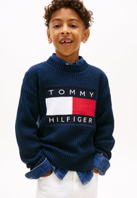 Σκουρόχρωμο πλεκτό πουλόβερ με ψηλό γιακά, που φέρει ένα λογότυπο με κόκκινα και λευκά τετράγωνα. Από κάτω διακρίνεται ένα μπλε τζιν πουκάμισο.