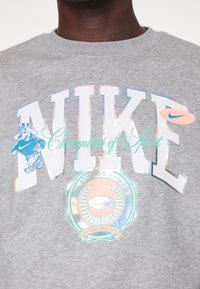 Grå sweatshirt med stor reflekterande "NIKE"-text, en logotyp med en tecknad figur och en cirkulär emblem med en lagerkransdesign.