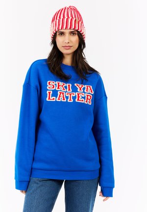 Blauwe trui met de tekst "SKI YA LATER" in rood en wit, met gestructureerde borduring. Gecombineerd met een rood en wit gestreepte gebreide beanie.