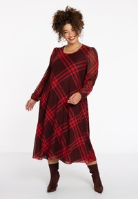 Rot-schwarz kariertes Kleid mit langen, transparenten Ärmeln, rundem Ausschnitt und fließender Silhouette, kombiniert mit braunen Stiefeln mit Absatz.