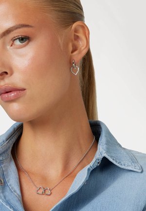 Orecchini a forma di cuore in argento con design pendente e una collana in argento con due cuori intrecciati, indossati su una camicia in denim.