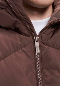 Smeđi puffer kaput s visokim ovratnikom, prednjim patentnim zatvaračem i teksturiranim materijalom. Sadrži oznaku s logotipom blizu patentnog zatvarača i bijelu unutarnju podstavu.