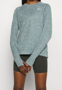Helle teal langärmlige Sportoberteil mit melierter Textur, tailliertem Schnitt und einem kleinen grauen Nike-Logo auf der linken Brust.