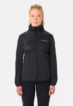 Donna con capelli biondi e ricci, indossa una giacca con cappuccio nera Vaude con cerniera, in piedi contro uno sfondo chiaro uniforme, rivolta in avanti.