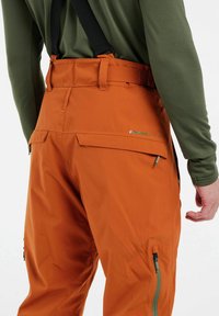 Oranje waterdichte broek met verstelbare bretels, zijzakken en ritsen. Materiaal lijkt glad met een duurzame textuur. Merkenlogo zichtbaar.