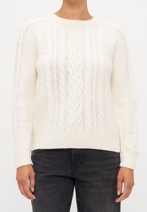 Woman wearing cream-colored cable knit sweater with long sleeves and ribbed cuffs, paired with dark gray jeans.