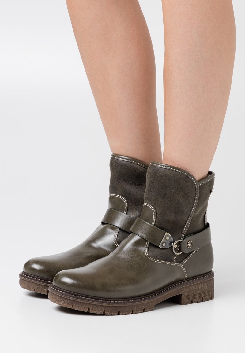 Bottes en cuir olive foncé montant à la cheville avec semelles en caoutchouc marron, sangles décoratives et coutures blanches, portées sur des jambes nues.