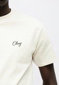 Άτομο που φοράει κοντομάνικη μπλούζα ανοιχτού μπεζ χρώματος με τη λέξη "Obey" σε μαύρο γραφικό κείμενο στο αριστερό μέρος του στήθους.