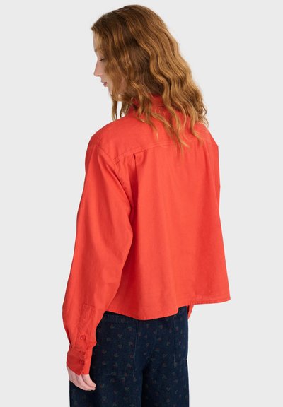 Femme aux cheveux roux ondulés portant une chemise rouge ample à manches longues et un pantalon bleu foncé à motifs, de dos sur un fond uni.