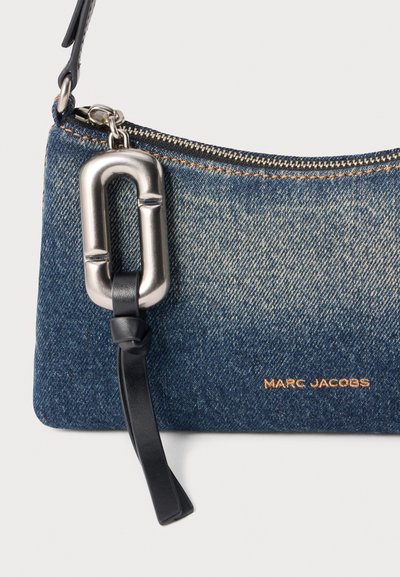 Pochette en denim Marc Jacobs avec tirette de fermeture éclair rectangulaire argentée et sangle en cuir noire sur fond clair.