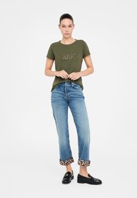 T-shirt verde oliva con scritta splendente "LIVIO." Abbinato a jeans in denim blu chiaro con orli a stampa leopardata e mocassini neri.