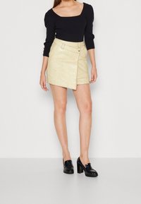 Schwarzes, geripptes Langarmshirt mit quadratischem Ausschnitt, kombiniert mit einem knielangen, metallic-goldenen Wickelrock mit seitlicher Verschlussnaht. Schwarze Loafers mit Absatz.