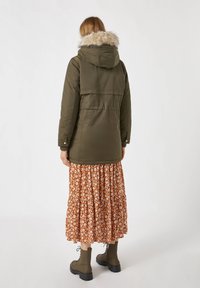 Olijfgroene parka-jas met een met bont gevoerde capuchon, voorzien van drukknoopsluitingen en een losse pasvorm, gecombineerd met een bruine maxi-jurk met bloemenprint.