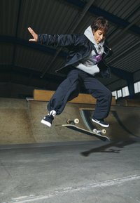 Skater w czarnych spodniach i kurtce, szarej bluzie, wykonujący trik. Powierzchnia to gładki asfalt z quarter pipe w tle.