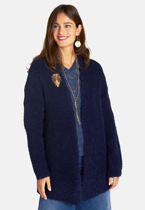 Navy-blaues, flauschiges Cardigan mit offenem Vorderteil, langen Ärmeln und lockerer Passform. Akzentuiert mit einer goldenen Blattbrosche und geschichteten Halsketten.
