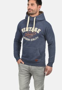 Blauer Hoodie mit einer Kängurutasche, cremefarbenen Kordeln und vintage-stiliger gedruckter Schrift in Orange und Weiß. Texturiertes Material mit gerippten Bündchen.