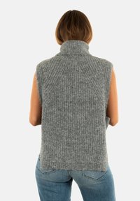 Donna che indossa un maglione senza maniche grigio a collo alto e jeans blu, vista da dietro su uno sfondo semplice.