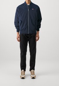 Námořnická bomber bunda se zapínáním na přední zip, elastickými manžetami a bočními kapsami. Nasažená přes černé džíny, doplněná světle hnědými teniskami.