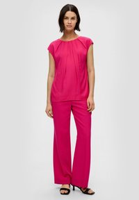 Top fucsia con dettagli plissettati, abbinato a pantaloni larghi fucsia. Entrambi realizzati in tessuto morbido; la modella indossa sandali neri con tacco.