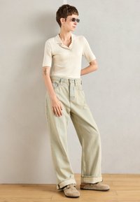 Personne portant un polo à manches courtes côtelé blanc cassé, un jean large léger, des chaussures beige à enfiler et de petites lunettes de soleil rectangulaires contre un mur uni.