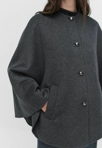 Manteau en laine grise avec un col arrondi, des poches latérales et des boutons noirs. Il possède des manches amples et un ourlet droit.