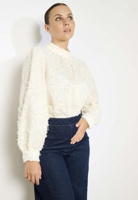 Blusa bianca con tessuto texturizzato e volant, colletto alto e maniche a sbuffo. Abbinata a jeans in denim scuro a vita alta.
