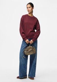 Mulher vestindo um sweatshirt bordeaux e jeans de perna larga azuis, segurando uma bolsa com padrão de leopardo, de pé contra um fundo simples.