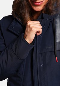 Veste rembourrée bleu marine avec une fermeture éclair sur le devant, des fermetures à pression et une capuche. Dotée d'un patch texturé sur l'épaule et de poignets élastiques avec logo.