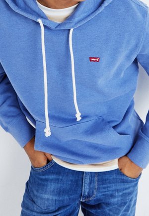 Personne portant un sweat à capuche Levi's bleu clair avec des cordons blancs et un jean bleu, les mains dans les poches, sur un fond uni.