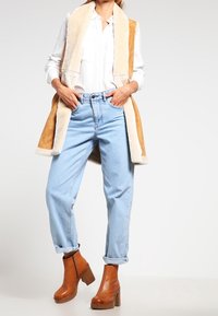 Tan faux fur-väst med beige detaljer, vit skjorta, ljusblå högmidjade jeans och bruna klackstövlar.