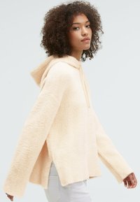Light peach, fuzzy hoodie with a loose fit, long sleeves, and a slight split at the sides. Features a drawstring at the neckline.