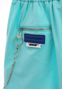 Una prenda turquesa que presenta un bolsillo con cremallera con una cadena plateada y una etiqueta rectangular azul con texto, destacando elementos de diseño funcional.