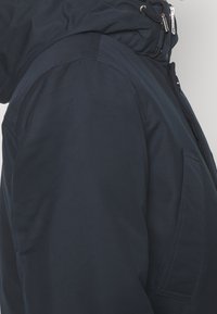 Laivastonsininen hupullinen takki, jossa on sileä pinta, vetoketjullinen etuosa ja sivutasku; siinä on jämäkkä siluetti ja pelkistetty muotoilu.