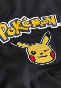Juodas audinys su išsiuvinėtu Pikachu veidu, puoštu geltonomis ir raudonomis detalėmis, šalia ryškiu geltonu ir mėlynu šriftu pateikiamas "Pokémon" logotipas.
