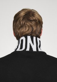 Černý pletený svetr s vysokým límcem, který má výrazný bílý vzor s logem. Textura je hladká a design je sportovní a moderní.