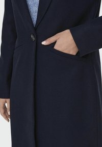 Cappotto di lana navy con chiusura a un bottone, tasche laterali e colletto a punta. Il tessuto ha una consistenza liscia e linee pulite.