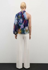 Femme portant un chignon blond, un pantalon blanc à jambes larges et un haut coloré à une manche style dos nu, debout contre un mur uni.